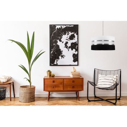 Závesná lampa svieti v interiéri. Lampa je valcového tvaru, čierna s dvoma striebornými pruhmi. Nachádza sa nad drevenou komodou v obývacej izbe s rastlinou a kreslom.