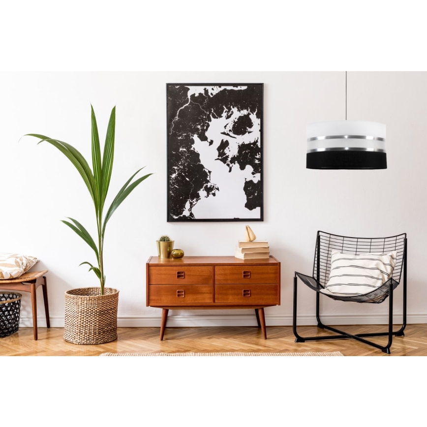 Závesná lampa svieti v interiéri. Lampa je valcového tvaru, čierna s dvoma striebornými pruhmi. Nachádza sa nad drevenou komodou v obývacej izbe s rastlinou a kreslom.