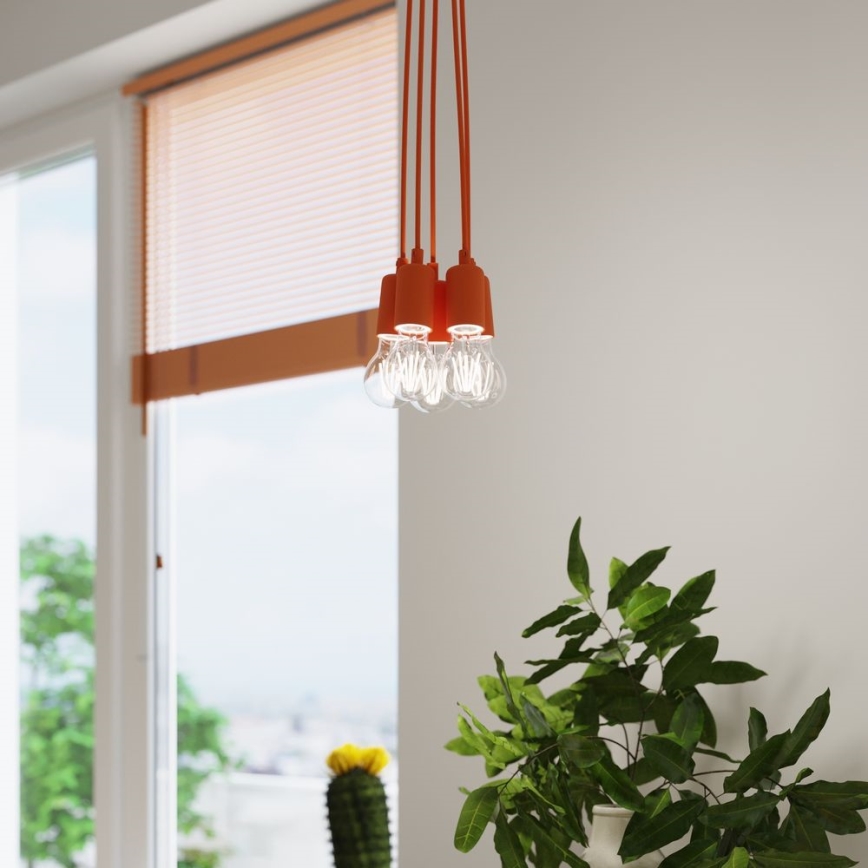 Oranžové svietidlo s viacerými žiarovkami visí v miestnosti pri okne so žalúziami. Rastlina v kvetináči je v popredí.