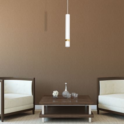 Visí lampa, cylindrického tvaru s bielym telom a zlatým pásikom, v hnedo-sivej miestnosti. Pred hnedým konferenčným stolíkom stoja dva biele kreslá s tmavohnedými rámami.