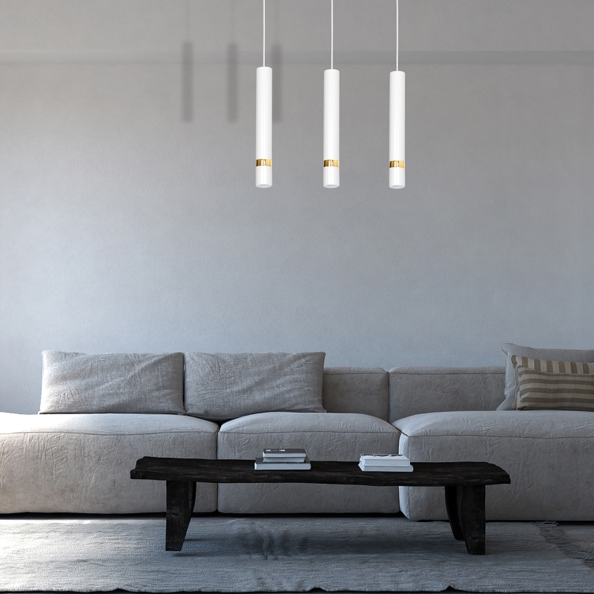 Tri visiace lampy, valcovitého tvaru s bielym tienidlom a zlatým pásikom, osvetľujú sivú sedačku s nízkym konferenčným stolíkom v svetlej miestnosti.