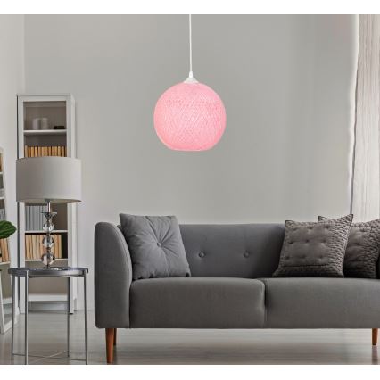 Ružové guľaté svietidlo visí zo stropu v obývacej izbe. Svietidlo je umiestnené nad sivou pohovkou s vankúšmi. V pozadí je vidieť knižnicu a stolík.