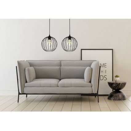 Dve čierne kovové závesné lampy s guľovým tienidlom visia nad svetlosivou pohovkou. V pozadí je rám obrazu s textom „ME HE DS“.