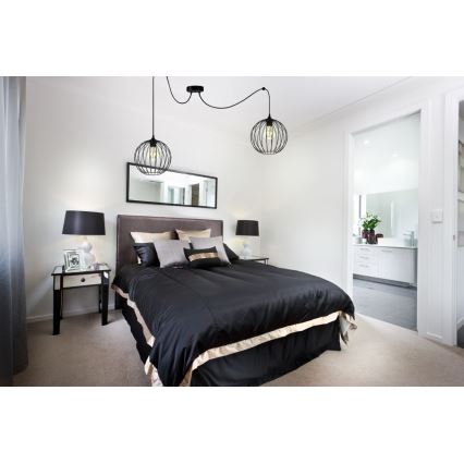 Dvojité závesné svietidlo s kovovými tienidlami v tvare gule svieti nad posteľou v spálni. Posteľ je prezlečená do čiernej prikrývky s béžovým lemom. Spálňa obsahuje aj nočné stolíky a zrkadlo.