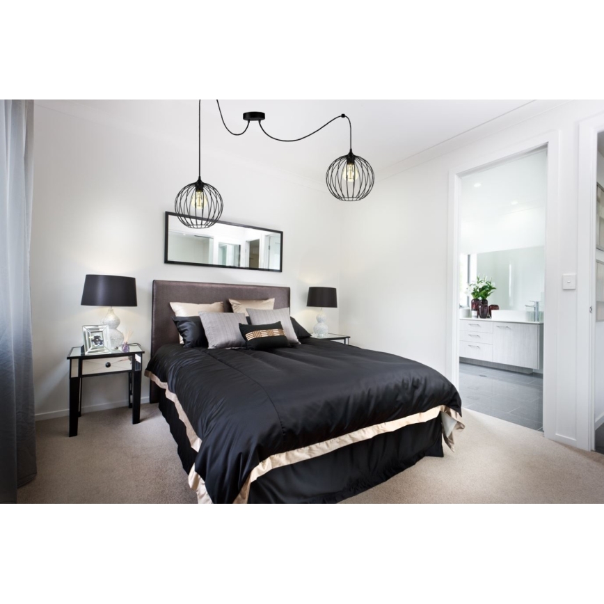Dvojité závesné svietidlo s kovovými tienidlami v tvare gule svieti nad posteľou v spálni. Posteľ je prezlečená do čiernej prikrývky s béžovým lemom. Spálňa obsahuje aj nočné stolíky a zrkadlo.