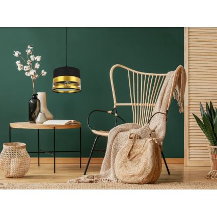 Závesná lampa visí v interiéri. Lampa má čierny a zlatý tienidlo. Nachádza sa nad nízkym stolíkom v miestnosti s ratanovým kreslom a doplnkami.