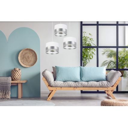 Tri visiace svietidlá s bielym a strieborným tienidlom osvetľujú obývaciu izbu. Svietidlá sú umiestnené nad drevenou pohovkou s bledomodrými vankúšmi. Pohovka stojí v priestore s rastlinami a svetlomodrými stenami.