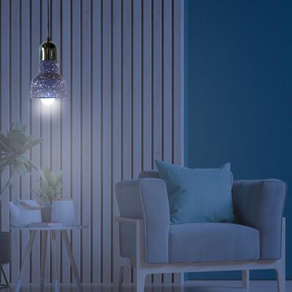 Závesné svietidlo svieti v interiéri. Svietidlo je tmavomodré s bielymi škvrnami, zlatým vrchnákom a visí v miestnosti s drevenou stenou a sivou pohovkou. Vedľa pohovky je malý stolík s kvetináčmi.