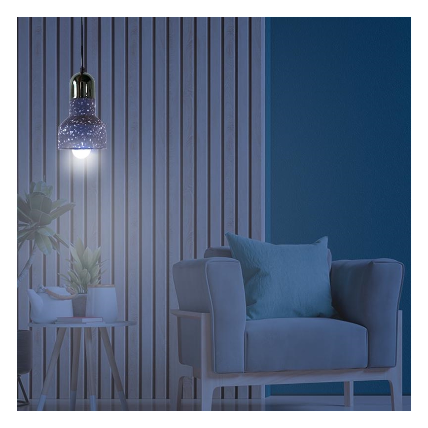 Závesné svietidlo svieti v interiéri. Svietidlo je tmavomodré s bielymi škvrnami, zlatým vrchnákom a visí v miestnosti s drevenou stenou a sivou pohovkou. Vedľa pohovky je malý stolík s kvetináčmi.