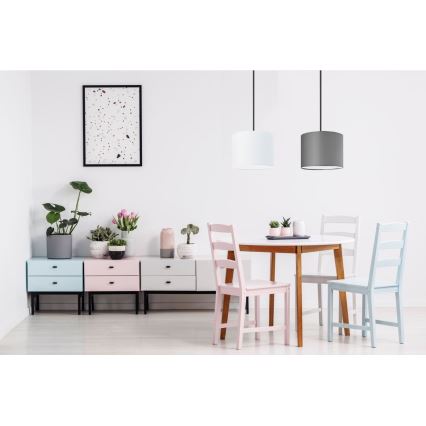 Závesné svietidlo svieti nad jedálenským stolom. Stolík je obklopený stoličkami v pastelových farbách. Za stolom sa nachádza nábytok s kvetináčmi a dekoráciami.