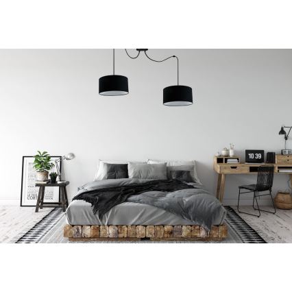 Dvojica čiernych textilných závesných lámp svieti nad posteľou v spálni. Lampa je pripevnená k stropu. V pozadí je vidieť drevený písací stôl a stoličku.