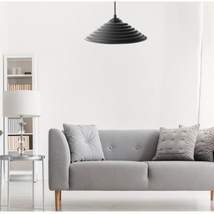 Čierna závesná lampa s ryhovaným tienidlom visí v svetlej obývacej izbe nad šedou pohovkou s vankúšmi. V pozadí je vidieť biely regál a stolík.