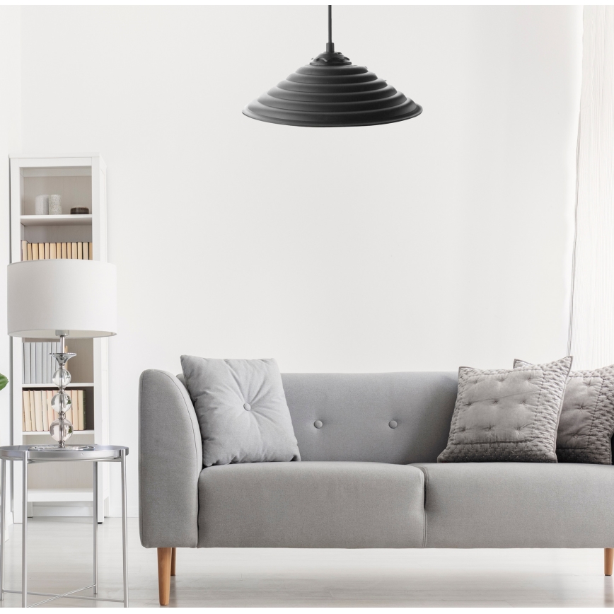 Čierna závesná lampa s ryhovaným tienidlom visí v svetlej obývacej izbe nad šedou pohovkou s vankúšmi. V pozadí je vidieť biely regál a stolík.