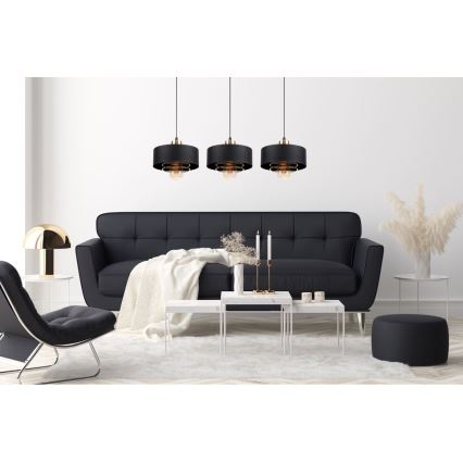 Tri čierne závesné lampy s mosadznými detailmi visia nad tmavosivou pohovkou v modernom interiéri. Lampy sú umiestnené symetricky. V miestnosti sa nachádza aj konferenčný stolík a kreslo.