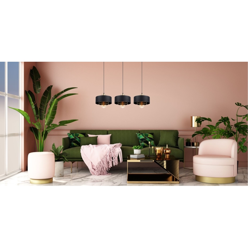 Tri čierne závesné lampy s mosadznými detailmi visia nad zelenou sedačkou v ružovej miestnosti. Sedačka je doplnená ružovým kreslom a doplnkami.