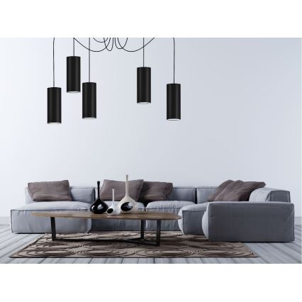 Päť čiernych valcových svietidiel visí z stropu nad sivou sedacou súpravou a nízkym dreveným stolíkom v svetlej miestnosti.