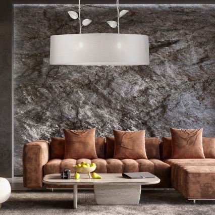 Dlhá obdĺžniková lampa s bielym tienidlom a kovovými listami visí v obývacej izbe. Svetlo svieti nad hnedou rohovou sedačkou a nízkym kávovým stolíkom s ovocím. Stena je z tmavého kamenného materiálu.