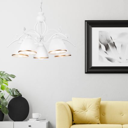 Biela päťramenná luster s kovovými ozdobami visí v svetlej miestnosti. Pod ním je žltá sedačka a vedľa je obraz v čiernom ráme.