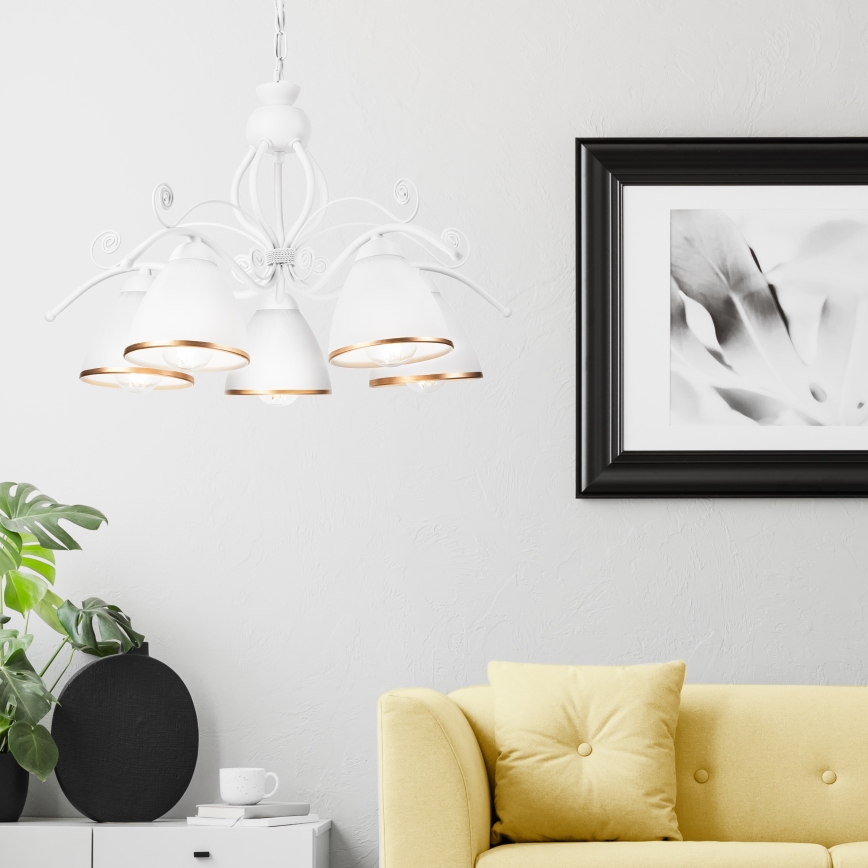 Biela päťramenná luster s kovovými ozdobami visí v svetlej miestnosti. Pod ním je žltá sedačka a vedľa je obraz v čiernom ráme.