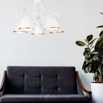 Biela lustrová lampa s piatimi tienidlami visí v miestnosti. Pod lampou je tmavosivá pohovka s drevenými podrúčkami. Vedľa pohovky stojí izbová rastlina v bielom kvetináči.