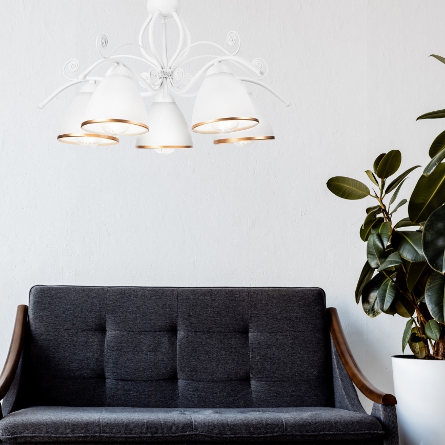 Biela lustrová lampa s piatimi tienidlami visí v miestnosti. Pod lampou je tmavosivá pohovka s drevenými podrúčkami. Vedľa pohovky stojí izbová rastlina v bielom kvetináči.