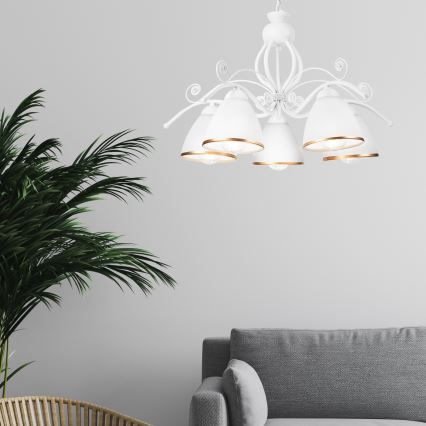 Biela lusterová lampa s piatimi tienidlami s pozláteným okrajom visí v miestnosti. Lampa je pripevnená k stropu. V pozadí je sivá pohovka a časť izbovej rastliny.