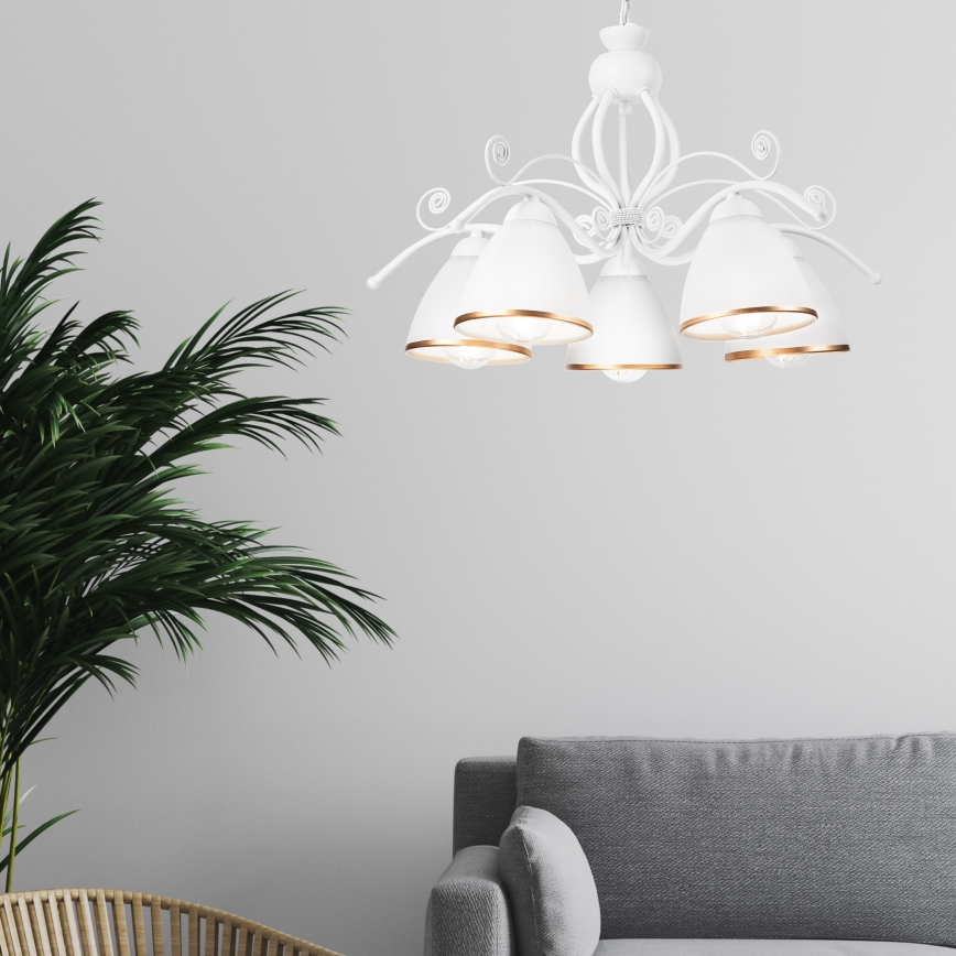 Biela lusterová lampa s piatimi tienidlami s pozláteným okrajom visí v miestnosti. Lampa je pripevnená k stropu. V pozadí je sivá pohovka a časť izbovej rastliny.