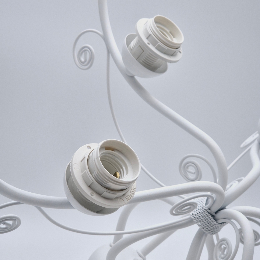 Biela kovová lampa s ozdobnými závitmi drží tri biele objímky na žiarovky. Objímky sú pripevnené k ramenám lampy. Svetlé pozadie.