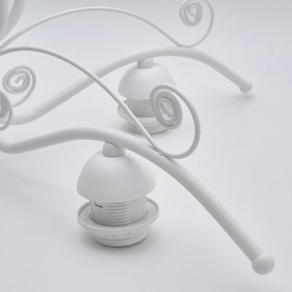 Biela kovová lampa s ozdobnými závitmi drží dve biele objímky na žiarovky. Lampa je položená na bielom podklade.