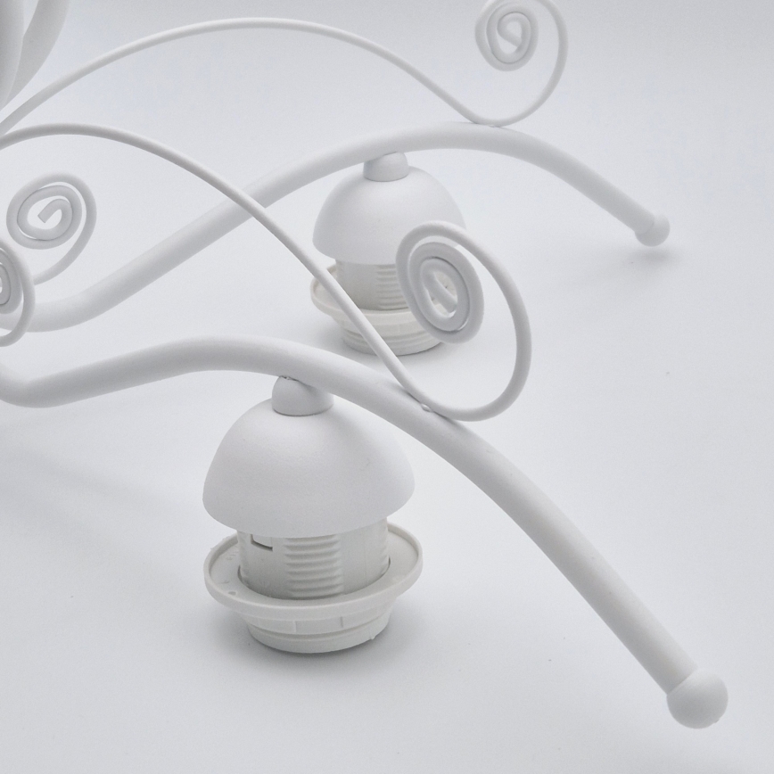 Biela kovová lampa s ozdobnými závitmi drží dve biele objímky na žiarovky. Lampa je položená na bielom podklade.