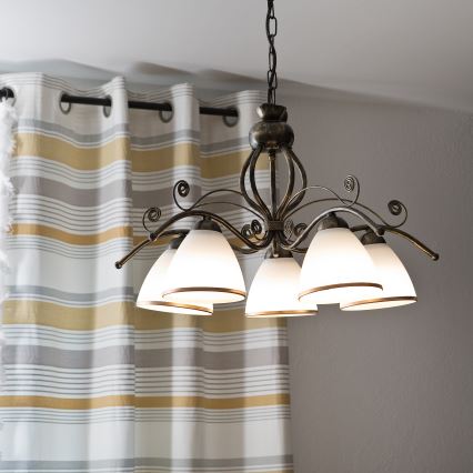 Lustr s piatimi tienidlami v tvare zvonu visí nad žltou pohovkou. Pohovka stojí v miestnosti s betónovou stenou. Na konferenčnom stolíku pred pohovkou sú knihy a váza s kvetmi.