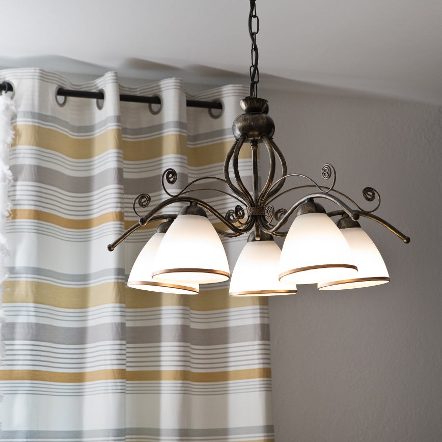 Lustr s piatimi tienidlami v tvare zvonu visí nad žltou pohovkou. Pohovka stojí v miestnosti s betónovou stenou. Na konferenčnom stolíku pred pohovkou sú knihy a váza s kvetmi.