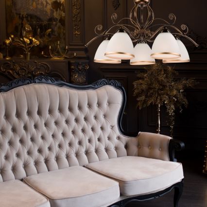 Čalúnená pohovka s čalúnením s prešívaním stojí v tmavej miestnosti. Nad ňou visí luster s viacerými tienidlami. V pozadí je tmavá stena s dekoratívnymi prvkami.