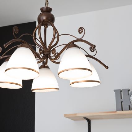 Kovová luster s viacerými tienidlami zo skla svieti v luxusne zariadenej miestnosti. Luster je umiestnený nad koženým kreslom a nízkym kávovým stolíkom. Interiér je štýlovo zariadený v hnedých a zlatých tónoch.