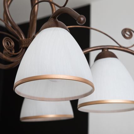 Bronzový luster s viacerými ramenami, zdobenými špirálami, je spojený ozdobným pleteným pásom. Ramenné zakončenia nesú mliečne tienidlá.