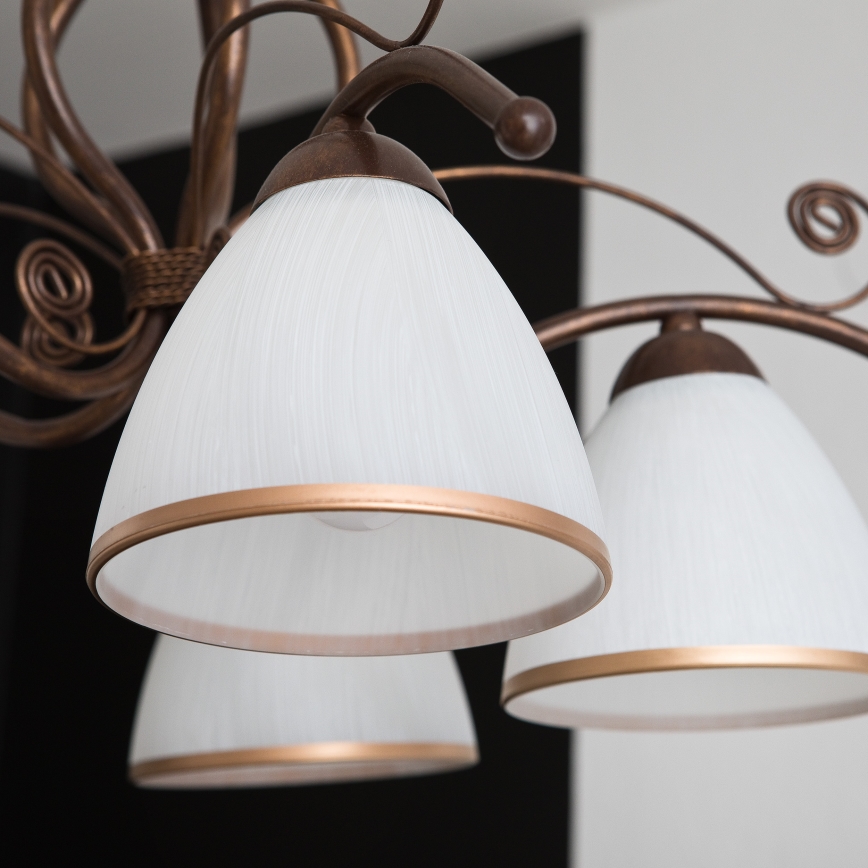 Bronzový luster s viacerými ramenami, zdobenými špirálami, je spojený ozdobným pleteným pásom. Ramenné zakončenia nesú mliečne tienidlá.