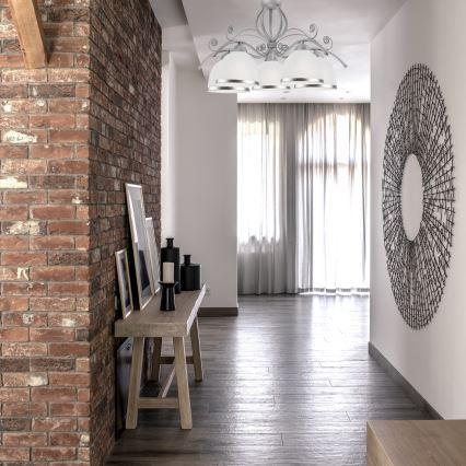 Drevený stolík s rámami a dekoráciami stojí pri tehlovej stene. Nad ním visí stropné svietidlo. V pozadí sa nachádzajú závesy a drevená podlaha.