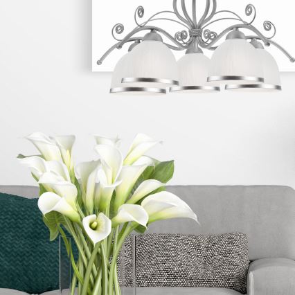 Stropné svietidlo s piatimi tienidlami z matného skla a kovovou konštrukciou visí nad šedou sedačkou. Kytica bielych kall žiari v priehľadnej váze na konferenčnom stolíku pred sedačkou.