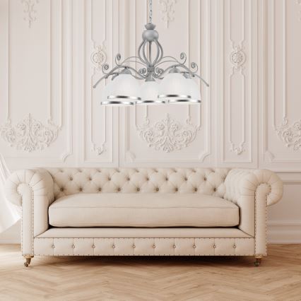 Stropná lampa s viacerými tienidlami visí nad pohovkou. Lampa je strieborno-šedá s bielymi tienidlami. Pohovka je svetlo béžová, čalúnená, v klasickom štýle. Nachádza sa v miestnosti s bielymi stenami a ozdobnými lištami.