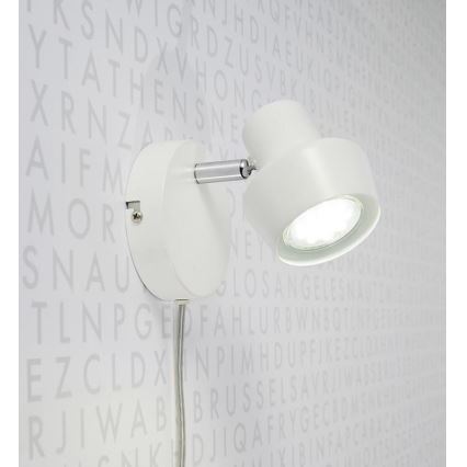 Biela nástenná lampa s otočnou hlavou svieti. Je pripevnená na stenu s pozadím z náhodne rozmiestnených písmen.