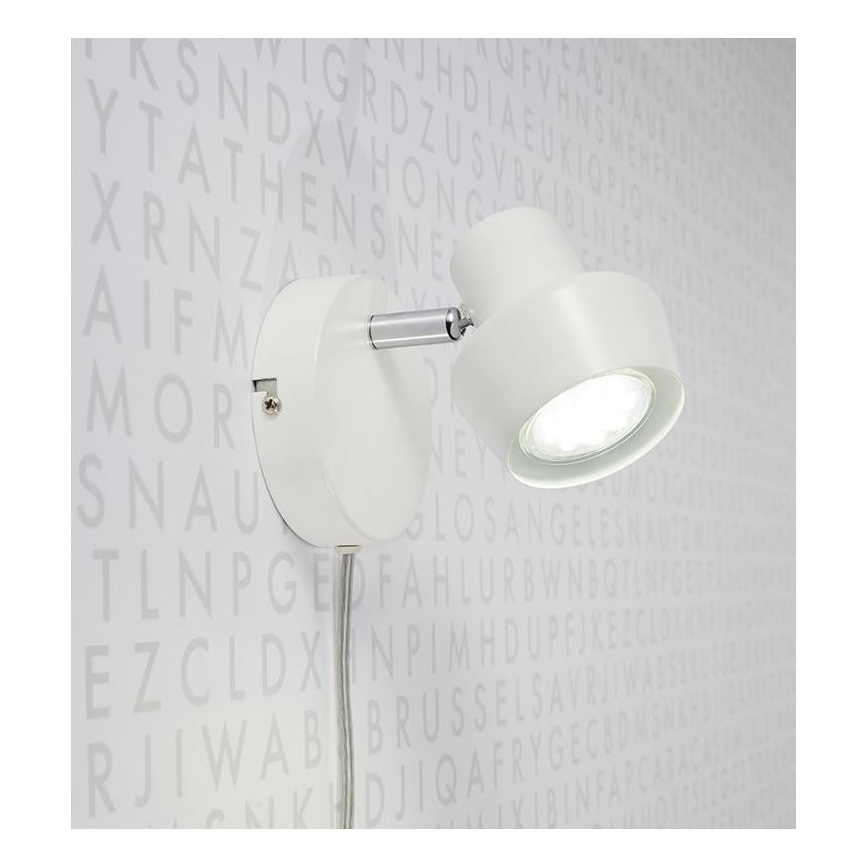 Biela nástenná lampa s otočnou hlavou svieti. Je pripevnená na stenu s pozadím z náhodne rozmiestnených písmen.