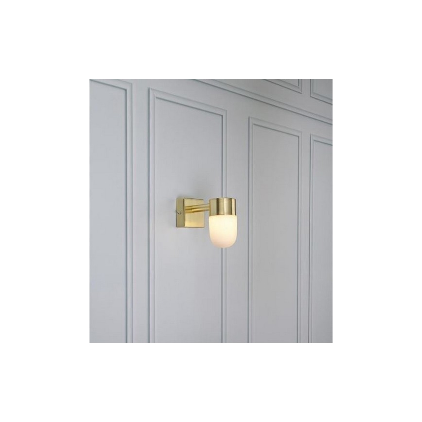 Zlatá nástenná lampa s matným skleneným tienidlom svieti na stene s panelovým obkladom.