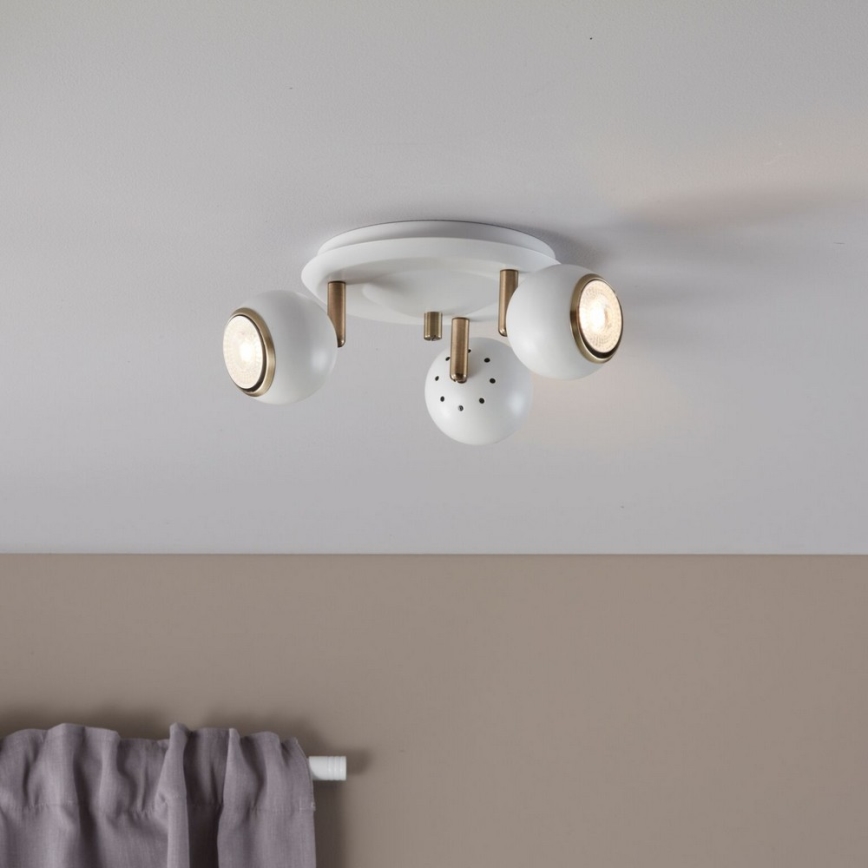 Trojramenné stropné svietidlo s bielymi guľovými tienidlami a zlatými detailmi je pripevnené k stropu. Svietidlo osvetľuje miestnosť. V popredí je časť steny a záves.