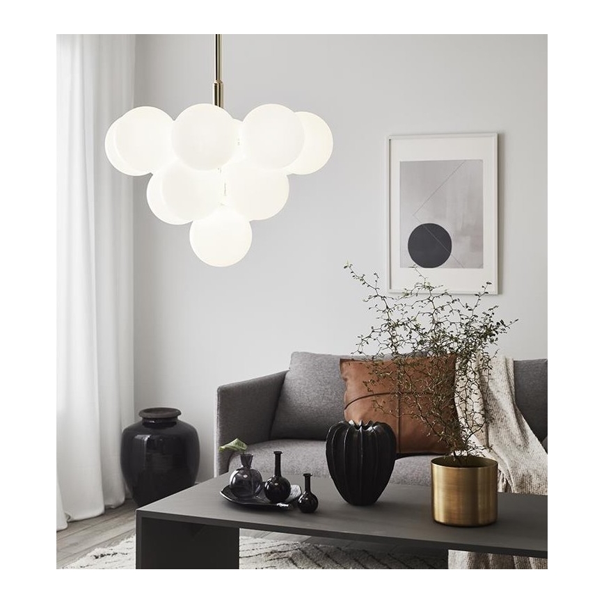 Lustr s viacerými bielymi sklenenými guľami visí v obývacej izbe. Na konferenčnom stolíku sú vázy a dekoračné predmety. Sivá pohovka s vankúšom a rastlina v zlatom kvetináči dopĺňajú priestor.