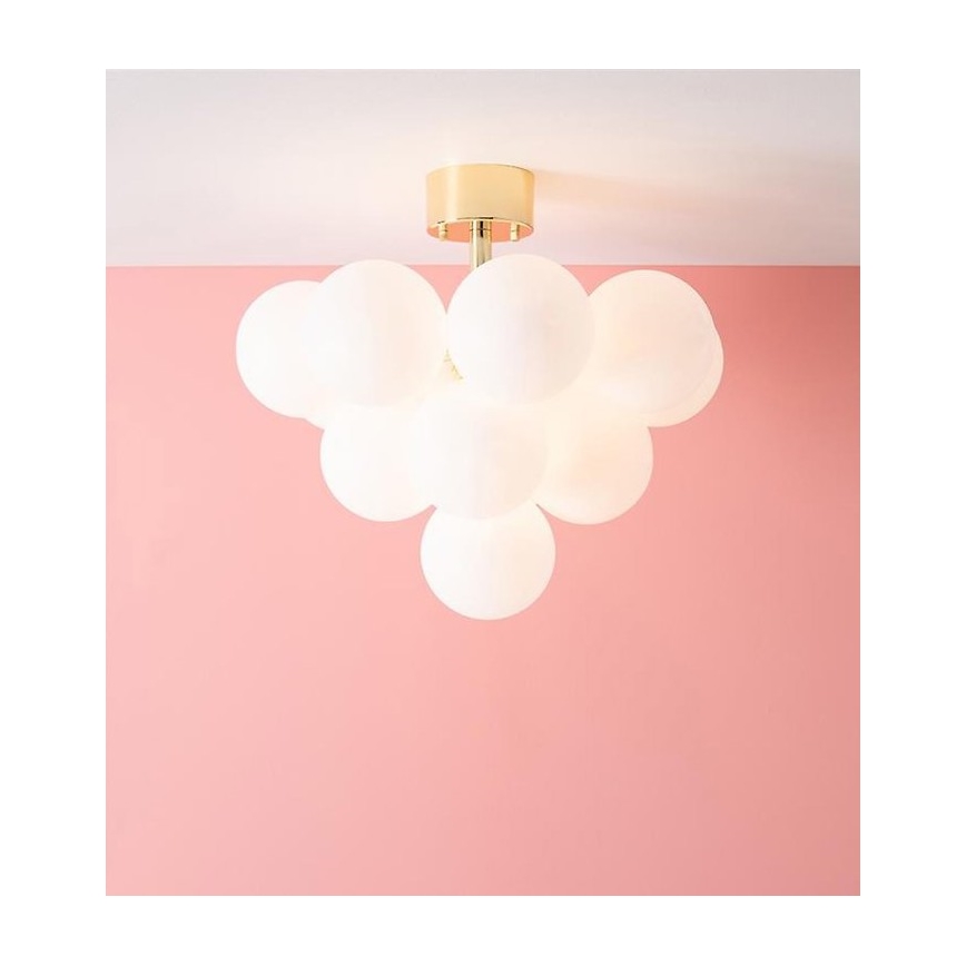 Závesné svietidlo s deviatimi bielymi guľovými tienidlami je pripevnené k stropu pomocou zlatej kovovej konštrukcie. Svietidlo je umiestnené v miestnosti s ružovými stenami.