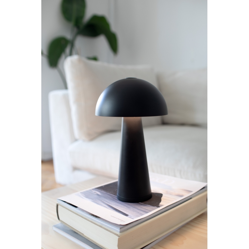 Čierna stolová lampa tvarom pripomínajúca hubu svieti na stolíku. Stolík je umiestnený pred svetlou pohovkou.