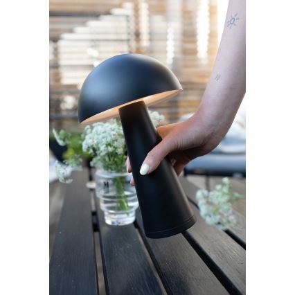 Čierna stolná lampa tvaru huby drží žena. Svieti. Stojí na tmavom drevenom stole, vedľa vázy s kvetmi.