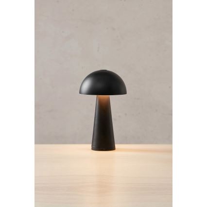 Stolová lampa svieti. Má čierny kupolovitý tienidlo a kužeľovitú základňu. Stojí na svetlom drevenom povrchu pred svetlohnedou stenou.