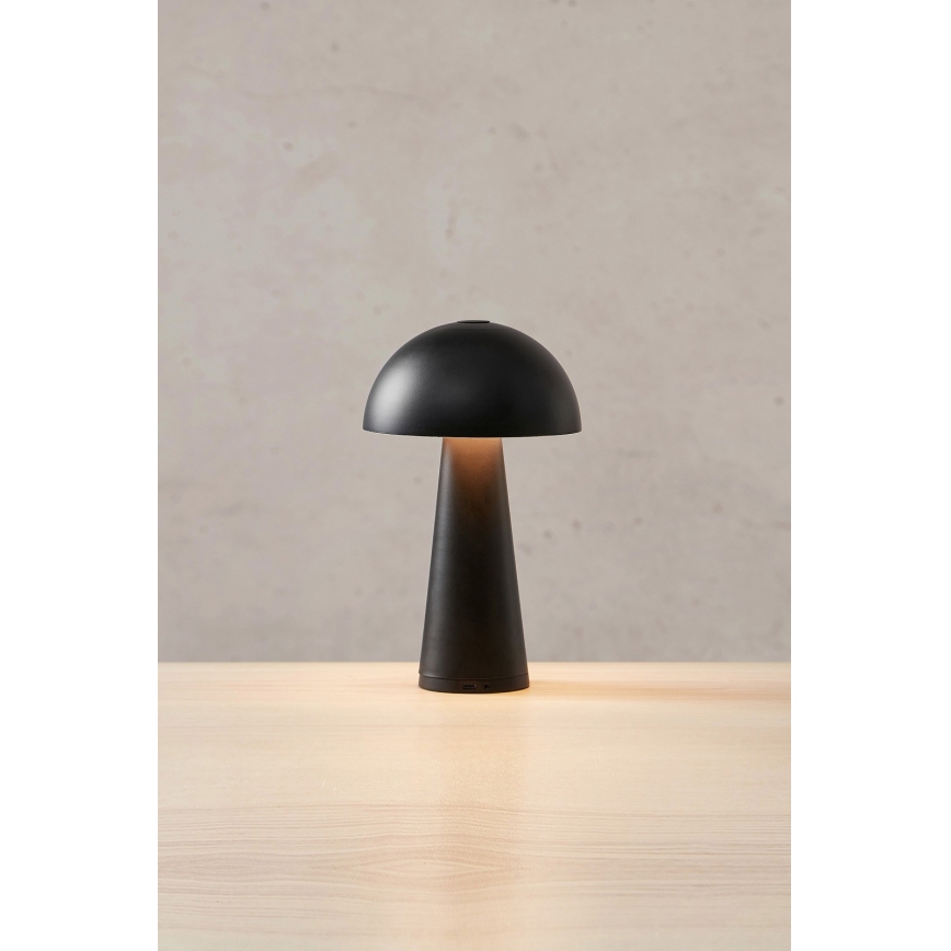 Stolová lampa svieti. Má čierny kupolovitý tienidlo a kužeľovitú základňu. Stojí na svetlom drevenom povrchu pred svetlohnedou stenou.