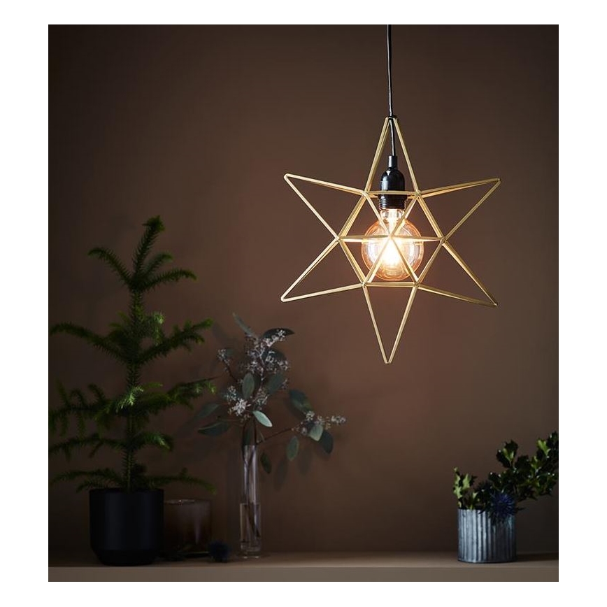 Závesné svietidlo v tvare hviezdy svieti. Svietidlo je zlaté, kovové a obsahuje žiarovku. V pozadí sú rastliny v kvetináčoch na poličke pred hnedou stenou.